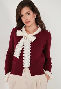 Burgundské pletené svetr s krémovým krajkovým límcem a mašlí. Má strukturované žebrované rukávy a krajkové detaily na manžetách.