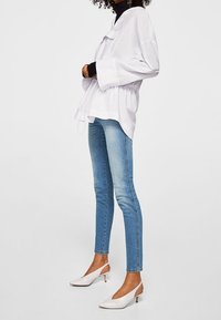 Camisa blanca holgada con cintura ajustable, llevada sobre una blusa negra; combinada con jeans skinny azul claro y tacones blancos tipo slingback.