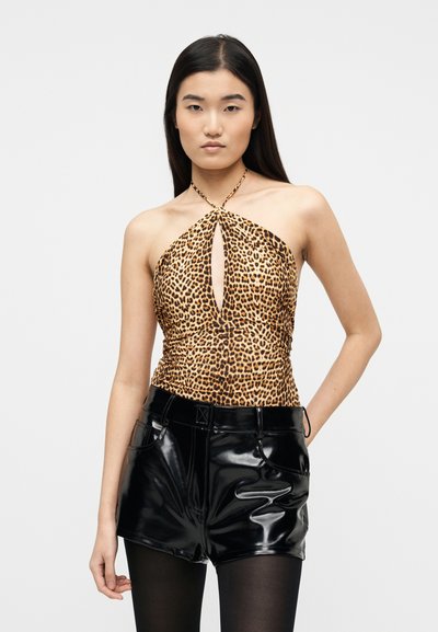 Jeune femme aux longs cheveux foncés portant un haut dos nu à imprimé léopard et un short en cuir noir brillant, posant devant un fond uni.