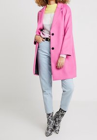 Manteau en laine rose à trois boutons, porté sur un pull rayé gris et jaune. Jean bleu clair et bottines à imprimé serpent noir et blanc.