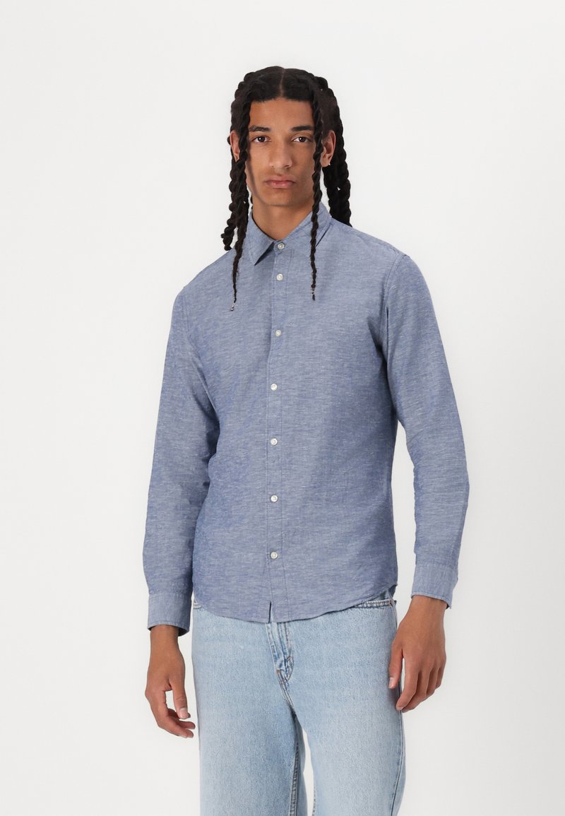 Camicia blu con bottoni realizzata in un tessuto testurizzato, dotata di colletto classico e maniche lunghe, abbinata a jeans in denim chiaro.