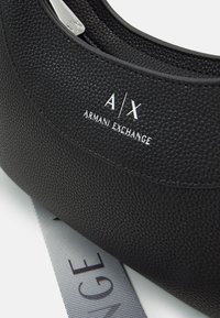 Czarna torebka z fakturowanej skóry od Armani Exchange z srebrnym logo i częściowo widoczną metką rozmiaru pod nią na białej powierzchni.