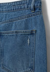 Izbleđeni plavi traperice s vidljivim šavovima, stražnjim džepom s vezom u obliku sivog munje, i oznakom marke na pojasu.