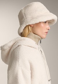 Bijeli krzneni šešir u obliku kante s teksturiranom, mekom površinom; uparen s svijetlim bež, visokim topom s patentnim zatvaračem i detaljem vezice.