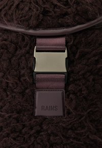 Tecido castanho texturizado com um acabamento peludo, apresentando um fecho metálico e uma etiqueta sólida escura em relevo com a inscrição "RAINS" na lateral.