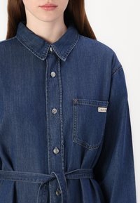 Abito di denim con colletto, chiusura frontale con bottoni, maniche lunghe e cintura in vita. Presenta una tasca sul petto con etichetta logata e trama blu.