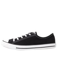 Zapatilla de lona negra con cordones blancos, puntera de goma y costuras blancas contrastantes. Presenta una suela plana con acentos de rayas negras.