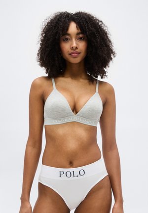 Femeie cu păr creț, purtând un sutien Polo Ralph Lauren gri deschis și chiloți Polo cu talie înaltă albi, stând în picioare pe un fundal simplu.