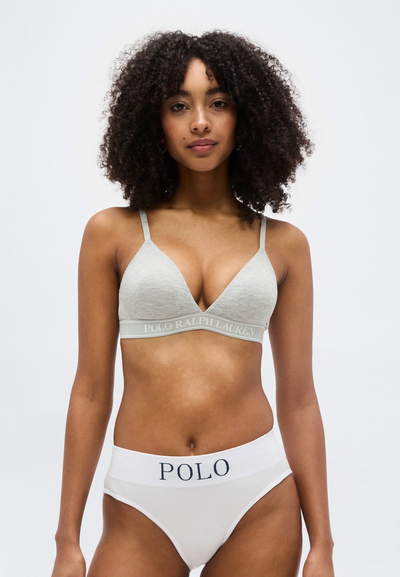 Γυναίκα με σγουρά μαλλιά, φορώντας ελαφρώς γκρι σουτιέν Polo Ralph Lauren και λευκά σλιπ Polo με ψηλή μέση, στέκεται μπροστά σε απλό φόντο.