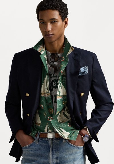 Marineblauwe double-breasted blazer met gouden knopen over een gemusterd groen shirt met boten. Gecombineerd met denim jeans en een bruine riem.