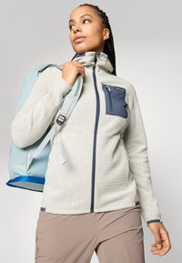 Hellgraue Fleece-Jacke mit Reißverschluss und strukturiertem Zickzackmuster, marineblaue Akzente und einer Brusttasche. Hellblauer Rucksack ausgestellt.