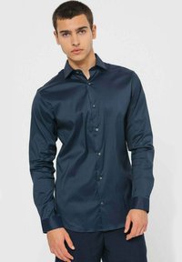 Camicia a maniche lunghe blu navy realizzata in un tessuto liscio, con chiusura frontale a bottoni e polsini a contrasto, indossata da un modello.