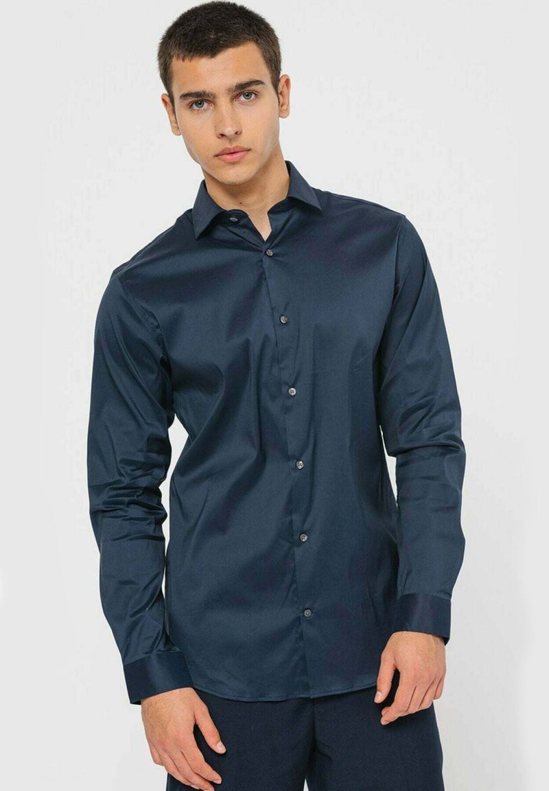 Camicia a maniche lunghe blu navy realizzata in un tessuto liscio, con chiusura frontale a bottoni e polsini a contrasto, indossata da un modello.
