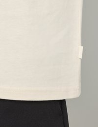 T-shirt en coton crème avec ourlet droit et étiquette latérale, associé à un pantalon en tissu texturé sombre. Design minimaliste, texture lisse.