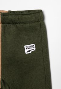 Pantalones deportivos verdes con una textura suave, con una franja lateral beige y un parche del logo de Puma en color blanco en la zona del muslo.