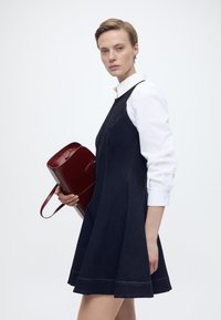 Γυναίκα με κοντά μαλλιά που φοράει σκοτεινό, αμάνικο φόρεμα πάνω από λευκό πουκάμισο με γιακά, κρατώντας μια μεγάλη σκούρα κόκκινη τσάντα, μπροστά σε απλό φόντο.