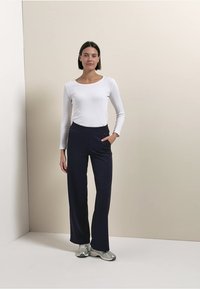 Weißes langärmliges Oberteil kombiniert mit marineblauen, weit geschnittenen Hosen. Die Hosen haben Seitentaschen und eine glatte Textur. Das Modell trägt silberne Turnschuhe.