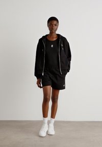 Svart zip-up hoodie över en svart linne och matchande shorts, kompletterad med vita strumpor och sneakers. Enkelt och sportigt design.
