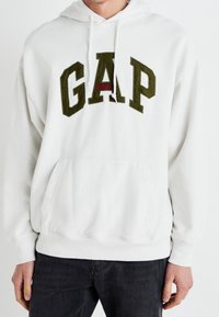 Bílá mikina s kapucí s velkým zeleným logem „GAP“ a červeným akcentem, vybavená klokánkou a stahovací kapucí. Bavlněná směs, ležérní střih.