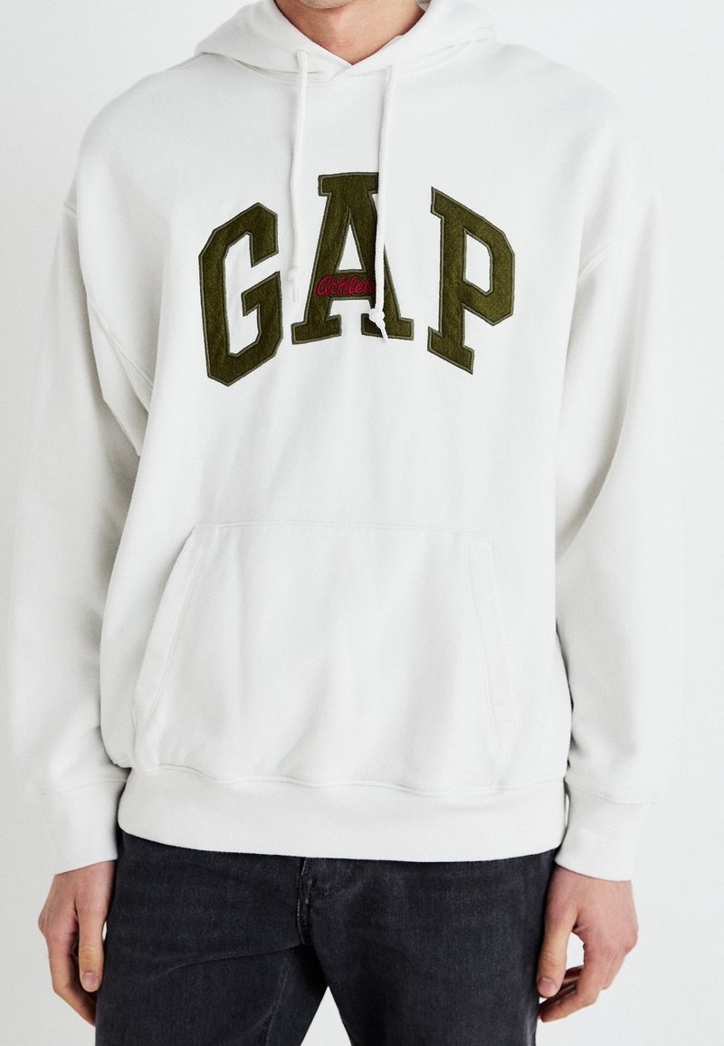 Bílá mikina s kapucí s velkým zeleným logem „GAP“ a červeným akcentem, vybavená klokánkou a stahovací kapucí. Bavlněná směs, ležérní střih.