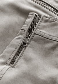 Gros plan d'une poche de veste en daim gris avec une fermeture éclair métallique noire partiellement ouverte et des détails de couture visibles.