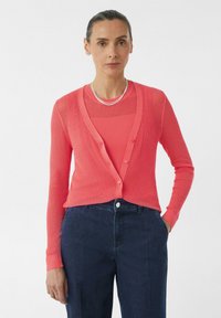 Korallfarbene, gerippte Strickjacke mit V-Ausschnitt, ausgestattet mit Knöpfen vorne und langen Ärmeln, kombiniert mit einem passenden texturierten Oberteil und dunklen Jeans.