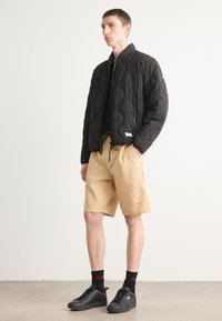Black quilted jacket with a high collar and zipper, paired with beige textured shorts. Model wears black sneakers and socks with red accents.