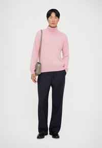 Lys rosa turtleneck genser, avslappet passform. Marineblå pinstripe bukser. Svarte sko. Liten grå skulderveske med flat design. Ensfarget bakgrunn.