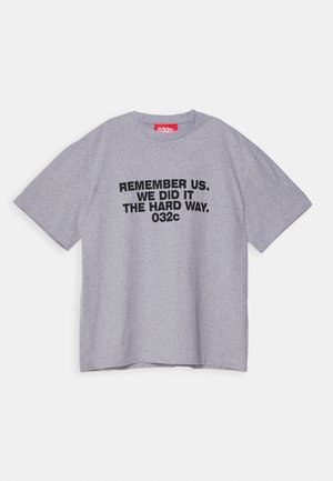 Szara koszulka z krótkim rękawem z czarnym napisem "REMEMBER US. WE DID IT THE HARD WAY. 032c" i czerwoną metką wewnątrz kołnierza.