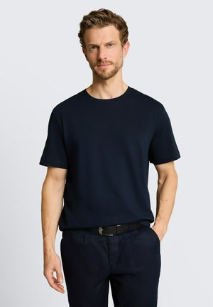 Mann mit kurzen lockigen Haaren, trägt ein schwarzes kurzärmeliges Hemd und schwarze Hose, eine Hand in der Tasche, vor einem einfarbigen hellen Hintergrund.
