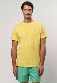 Kollane puuvillane T-särk, millel on ümar kaelus ja väike türkiissinine logo rinnal, koos piparmündiroheliste lühikesed pükstega.