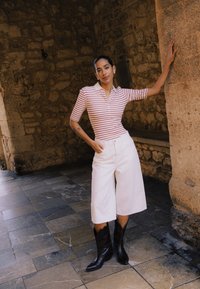 Femme portant une chemise rayée rouge et blanche, un short large blanc et des bottes noires, appuyée contre un pilier en pierre dans un cadre rustique.
