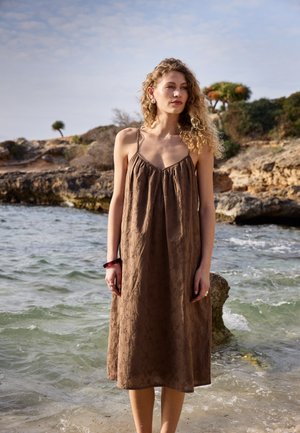 Femme aux cheveux bouclés portant une robe marron sans manches, debout dans l'eau peu profonde de l'océan près d'un rivage rocheux sous un ciel partiellement nuageux.