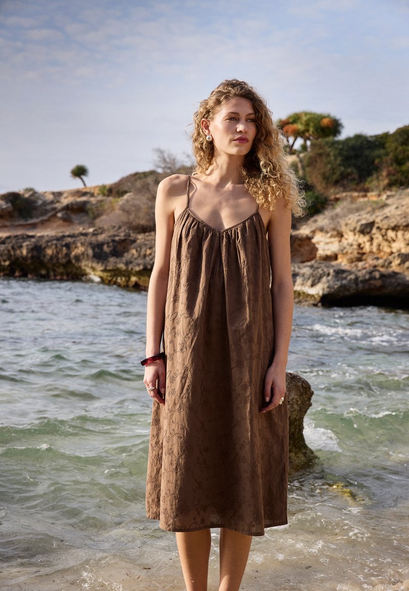 Femme aux cheveux bouclés portant une robe marron sans manches, debout dans l'eau peu profonde de l'océan près d'un rivage rocheux sous un ciel partiellement nuageux.