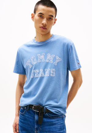 Modrá bavlnená tričko s nápisom "TOMMY JEANS" vytlačeným bielou farbou, krátke rukávy, okrúhly výstrih, nosené s tmavými denimovými džínsami a čiernym tkaným opaskom.