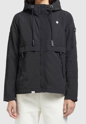 Veste noire à capuche avec fermeture éclair à l'avant, deux poches zippées, cordons et petit logo feuille d'érable blanche sur la poitrine, portée sur un pantalon blanc.
