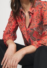 Blouse rouge à motifs avec un design floral, manches longues avec des poignets élastiques et un col à boutons. Le matériau semble léger et texturé.