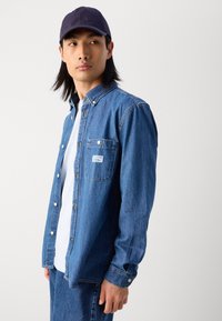 Denim shirt in medium blue with button-up front, chest pocket, and soft texture. Worn over a white t-shirt, paired with denim pants.