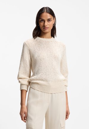 Vrouw met middellang donker haar die een crèmekleurige gebreide trui en lichtbeige broek draagt tegen een effen witte achtergrond.