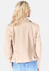 Beige leren motorjack met gestructureerde schouders, gedetailleerde stiksels en zijritsen, voorzien van een klein logo-embleem op de achterkant.