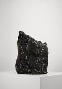 Zwarte en smaragdgroene handtas met slangenpatroon, bestaande uit zacht materiaal, een ontspannen vorm en een aangetrokken onderkant, tentoongesteld op een witte sokkel.