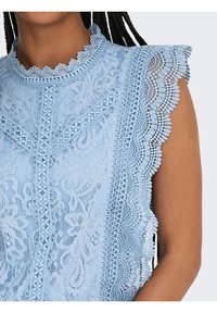 Haut sans manches en dentelle bleu clair avec un col montant festonné et des motifs floraux complexes porté par une personne aux cheveux tressés.