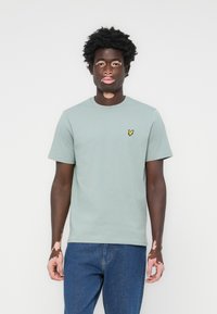 T-shirt en coton vert clair avec des manches courtes et un col rond. Présente un petit logo jaune brodé sur la poitrine gauche.