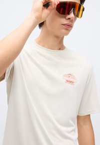 Νεαρός άνδρας που φοράει μπεζ μπλουζάκι Patagonia και ρυθμίζει μεγάλα αντανακλαστικά πορτοκαλί γυαλιά ηλίου, με απλό ανοιχτό φόντο.