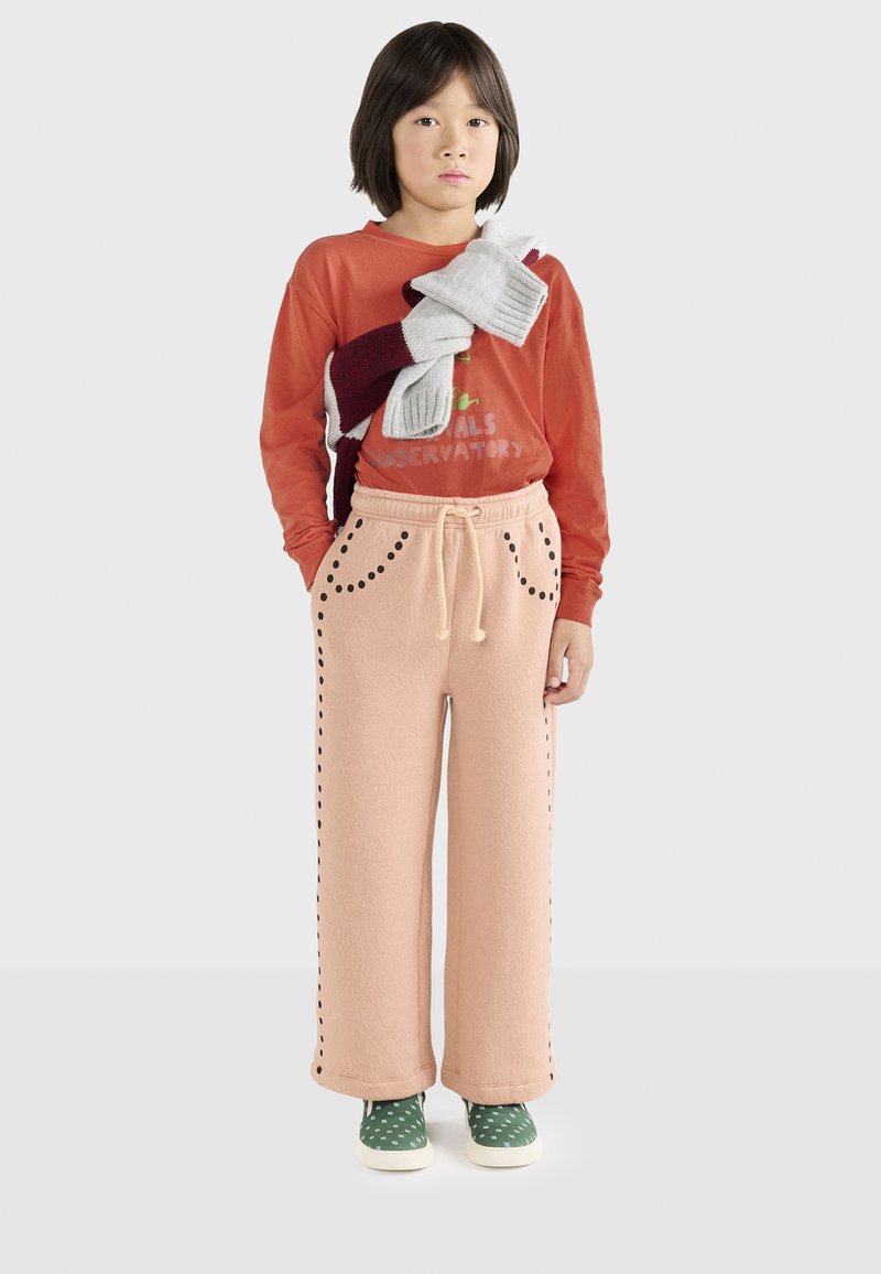 Pantaloni in pile rosa chiaro con dettagli di cucitura nera, abbinati a una maglietta a maniche lunghe arancione e a una miscela di foulard grigi e bordeaux drappeggiati sulla spalla. Sneakers verdi con pois bianchi.