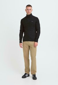Sort strikpullover med høj krave og lynlås, parret med tan lige ben bukser og sorte sneakers med hvide striber.