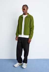 Green lightweight jacket with elastic cuffs, white t-shirt, black wide-leg pants, and silver athletic shoes with yellow accents.