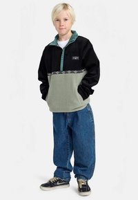 Pullover in pile nero e verde chiaro, con colletto testurizzato, patch logo sul petto e banda decorata. Abbinato a jeans larghi blu e sneakers.