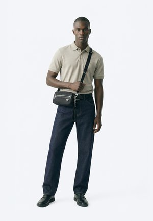 Mężczyzna w beżowej koszulce polo, ciemnych dżinsach, czarnych butach i czarnej torbie na ramię z zamkiem, stojący na jednolitym białym tle.