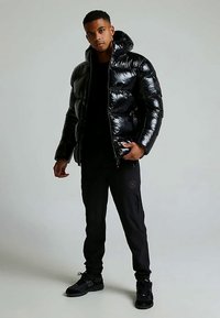 Svart dunjakke med blank finish, frontglidelås og hette. Brukes med svarte bukser, og viser et strømlinjeformet, moderne design.